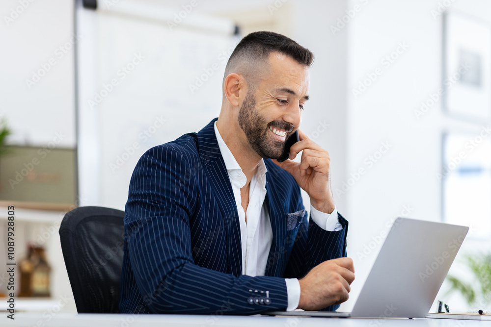 Poster a well-dressed man smiles as he has a phone conversation while working on his laptop in a bright, co
