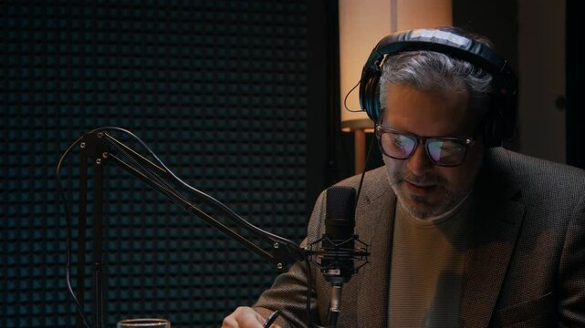 Medium shot of cheerful mature handsome Caucasian man in glasses, headphones talking into microphone while participating in podcast on controversial business or economy subjects