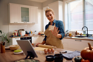 Happy woman having video call on touchpad while baking in kitchen.