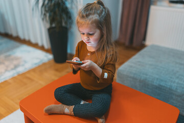 A little girl sits cross-legged on a small orange table, engrossed in a smartphone, depicting the...
