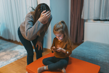 A woman showing concern as a child intently uses a smartphone indoors, highlighting modern family...