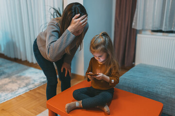 A woman stands distressed as a little girl is engrossed in her smartphone, symbolizing the...