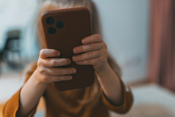 A pair of small hands belonging to a girl hold a smartphone upright, wrapped around the device. The home environment adds to the relaxed technological interaction.