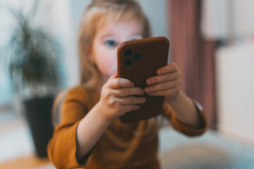 Fototapeta premium A young child holding a smartphone, deeply engaged, with a softly blurred background, highlighting concentration and modern technology interaction dynamics.