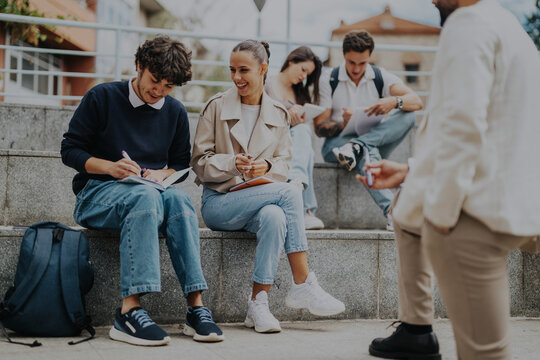 Group of high school students sitting outdoors, collaborating on tasks with the help of a professor, fostering teamwork and learning. Students are engaged in discussions and writing notes on a sunny