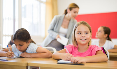 Obraz premium Portrait of cheerful blonde tween girl studying in classroom, listening to schoolteacher and writing in notebook