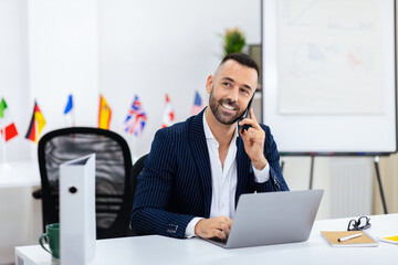 A man in a pinstripe suit smiles as he makes a phone call at his desk. A laptop and documents are present, with various country flags displayed in the background, suggesting an international setting.
