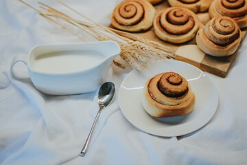 Fresh cinnabon on a plate on a background of other buns on a wooden tray and milk with a spoon 