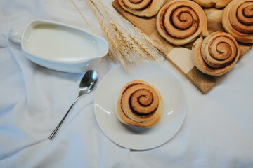 Fresh cinnabon on a plate on a background of other buns on a wooden tray and milk with a spoon 