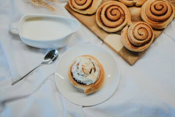 Fresh cinnabon on a plate on a background of other buns on a wooden tray and milk with a spoon