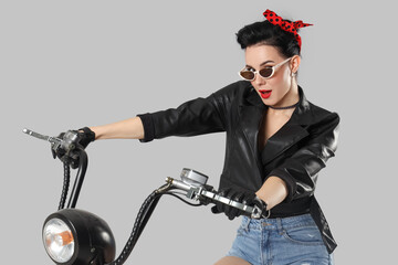 Young pin-up woman in sunglasses riding electric bike on light background