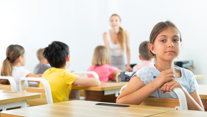 Smiling positive small school girl sitting at school desk in classroom on background with classmates