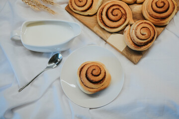 Fresh cinnabon on a plate on a background of other buns on a wooden tray and milk with a spoon 