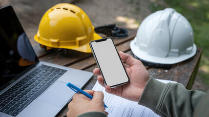 A person is holding a cell phone and a pen on a table. The person is wearing a yellow hard hat