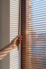 Hand adjusting wooden blinds to control sunlight in a bright room during morning