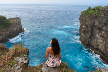 A beautiful woman in a pink dress sits on a cliff above the ocean on the island of Nusa Penida. Devil's Billabong an incredibly wonderful lagoon with splashes from the waves.