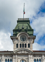 Trieste, Italy - June 26, 2024: Clock tower with statues ringing bell and flag on top of Historic City hall on on Piazza Unità d'Italia, Unity of Italy, Square, under blue cloudscape