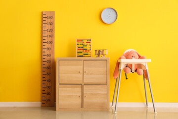 Toys on commode with wooden stadiometer and baby highchair near yellow wall in children's room