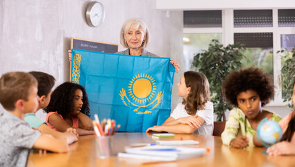 Fototapeta premium Friendly high school female teacher, conducting a lesson in the classroom, holds the national flag of Kazakhstan and tells .the pupils the history of the country