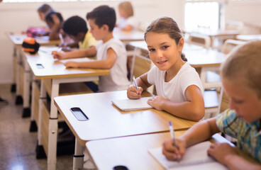 Focused girl sitting at desk writing test in classroom full of pupils during lesson