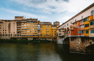 Ponte vecchio reflecting on arno river under cloudy sky with colorful buildings along the riverbank in florence, italy, creates a captivating urban landscape