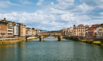 Fototapeta premium Ponte vecchio reflecting on arno river under a cloudy blue sky with historic buildings on both sides of the river in florence, italy, creating a picturesque cityscape