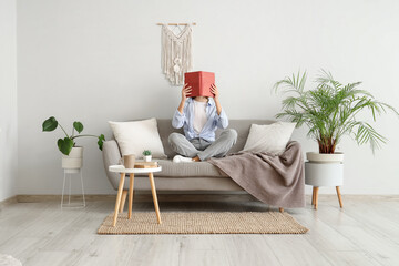 Pretty young woman reading book on sofa near white wall