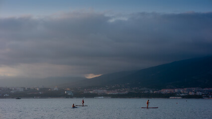 Obraz premium Paddleboarding at Sunset with Mountain View. Two people paddleboarding on calm water, picturesque coastal scene. Mountain backdrop, evening light, tranquil waters, outdoor recreation.