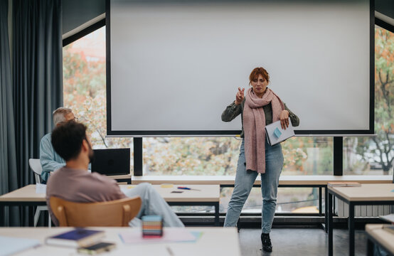A young woman stands confidently as she presents to students in a classroom, with an elderly professor observing. The scene captures an engaging learning atmosphere.