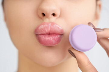 Beautiful young woman with lip balm blowing kiss on light background, closeup