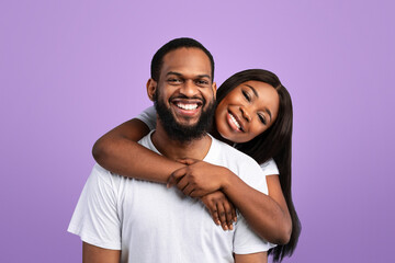 Naklejka premium Tenderness Concept. Portrait of charming cute black lady hugging her man from behind, enjoying time together, standing isolated over yblue studio background, posing and looking at camera