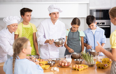 Young guy and adult woman cook in uniform teaches group of children how to cook dish