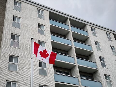 A Canadian flag waves in front of a white brick apartment building with blue balconies, highlighting the backdrop of the ongoing housing crisis in Canada. A symbol of pride amidst growing challenges