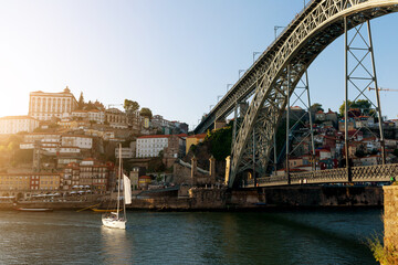 A sailboat moves gracefully on the calm waters beneath a striking bridge in Porto. The sunset casts...