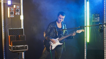 Obraz premium Independent iconic woman playing her electric guitar for a live performance in an edgy dark studio with spotlights. Confident powerful artist with a leather jacket and funky style. Camera B.