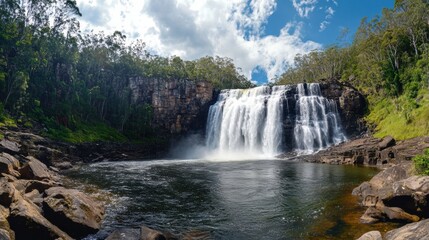 Fototapeta premium Majestic Waterfall Cascading Into a Serene Pool Surrounded by Lush Green Forest and Blue Sky, Capturing the Beauty of Nature in a Panoramic View