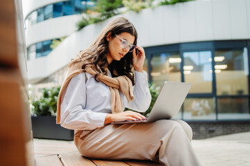 Young businesswoman working on laptop outside of modern business building	