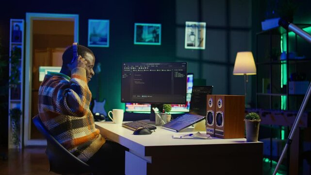 Black programmer remotely working in home office writing code on computer at his desk. Programming engineer IT expert in apartment typing lines of code at workstation, teleworking. Camera B.