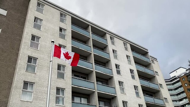 A Canadian flag waves in front of a white brick apartment building with blue balconies, highlighting the backdrop of the ongoing housing crisis in Canada. A symbol of pride amidst growing challenges