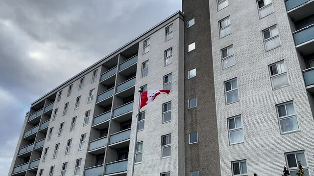 A Canadian flag waves in front of a white brick apartment building with blue balconies, highlighting the backdrop of the ongoing housing crisis in Canada. A symbol of pride amidst growing challenges