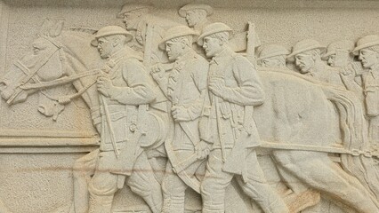 A detailed stone relief depicts WWI soldiers marching alongside a horse, carrying gear and weapons, commemorating military service in a solemn and timeless artistic representation.