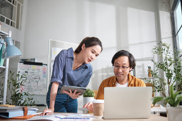 Two young Asian startup company partners discuss and brainstorm in casual small business office, marketing planning jobs on a laptop, and freelance work as E-commerce entrepreneur and coworker team.