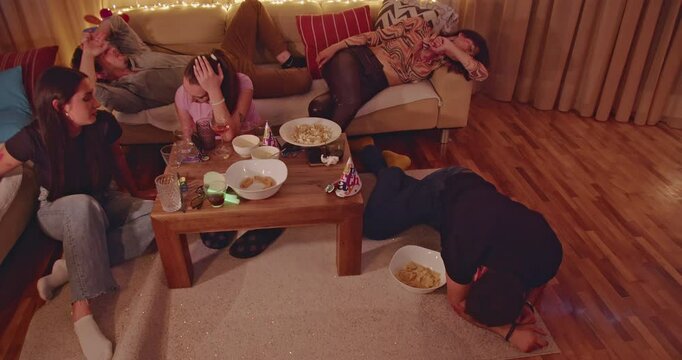 Friends passed out on couch and floor after a house party. Snacks and drinks remain on the coffee table. They are hungover.