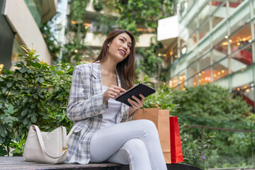 Asian Woman with Shopping Bags Using Digital Tablet Amid Greenery Cityscape Background