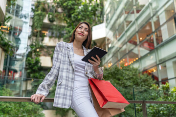 Asian Woman with Shopping Bags Using Digital Tablet Amid Greenery Cityscape Background