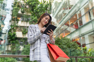 Fototapeta premium Asian Woman with Shopping Bags Using Digital Tablet Amid Greenery Cityscape Background