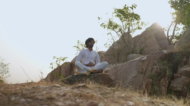 young Indian yogi doing his yoga mediation practices on hills rocks in forest