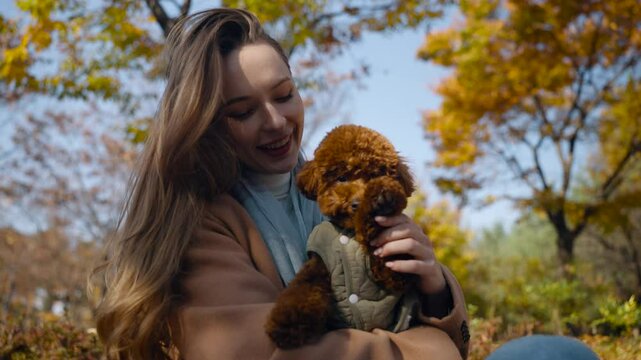 Smiling Woman Holding Toy Poodle Dog on Knees Waving Doggy's Paw Making bye-bye or Hello Gesture Sitting on Bench in Autumn City Park - dolly in