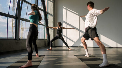 Yoga group. Balance exercise. Fit people practice warrior pose asana pilates lesson in activewear with female fitness instructor on sport mat in gym studio indoors.