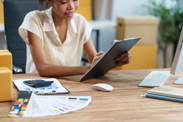 A beautiful African American woman sits at a desk, checking items, packing products, taking orders, monitoring sales on her computer. Packages are ready for shipping, showcasing her thriving online